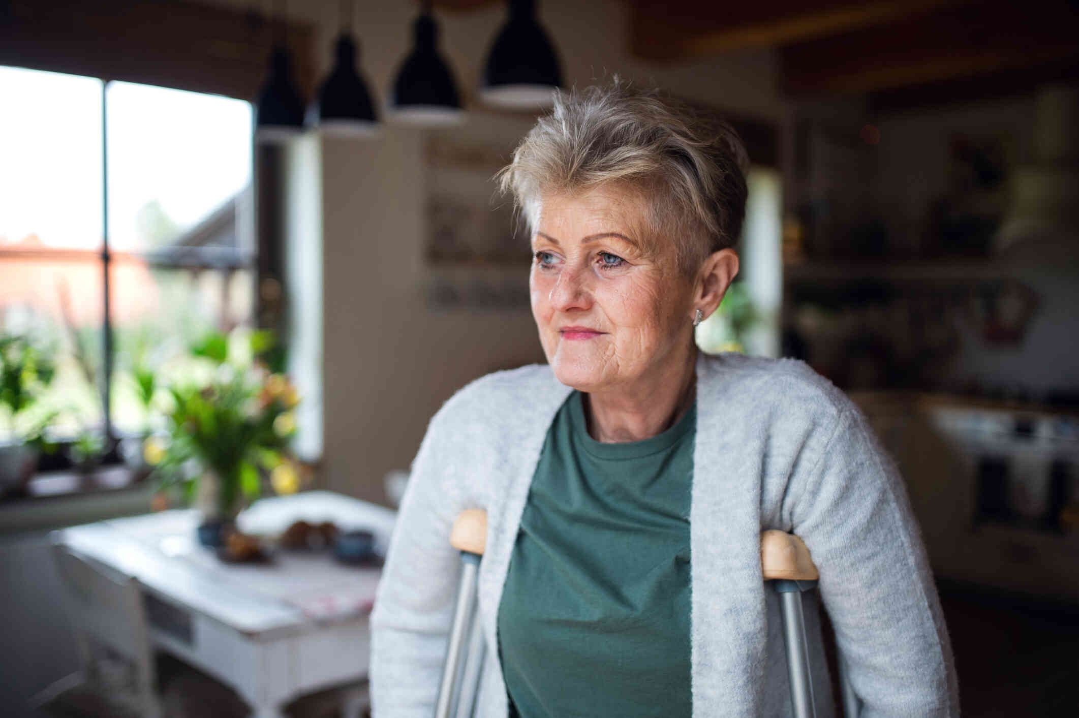 A close up of an elderly woman as she stands on crutches in her home with a sad expression.